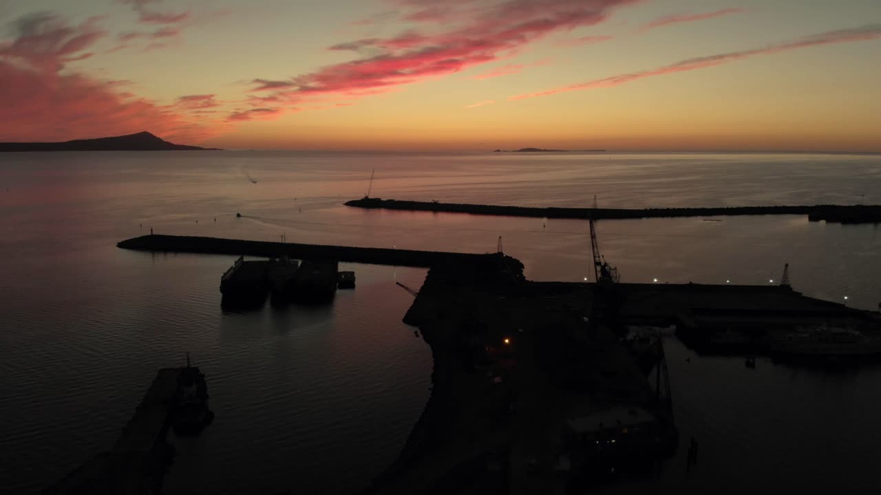 Veduta aerea con drone del porto al tramonto con mare calmo, cielo colorato e sagome di moli, paesaggio costiero con luce soffusa e atmosfera tranquilla sull'acqua.
