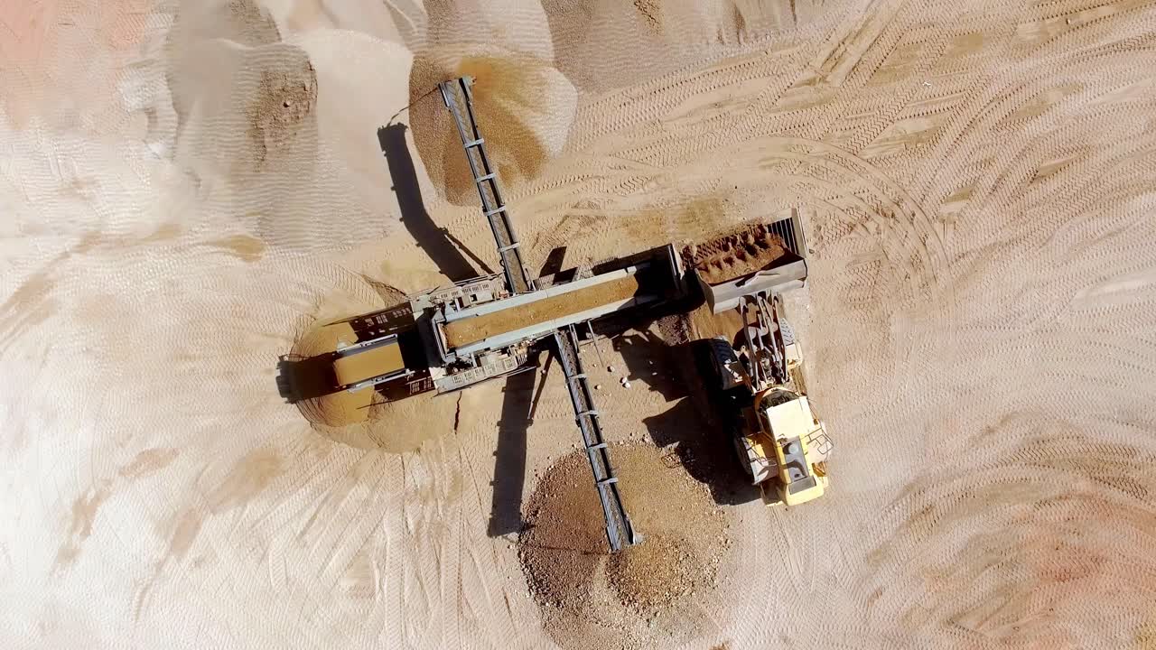 Front-end loader moving sand into a large screening plant that sorts the aggregate material into piles at a quarry or open-pit mine - A top-down aerial view