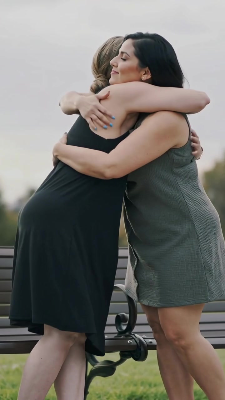 Video verticale: Donna in nero che si protende per abbracciare un'amica in abito verde oliva da una panchina del parco, salutando.