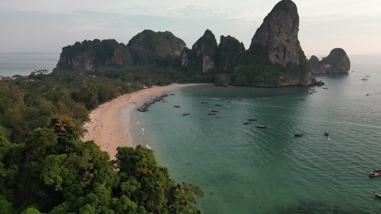 Panoramica aerea oltre un'imponente scogliera calcarea all'ora d'oro che rivela Railay West Beach a Krabi con barche longtail ancorate in acqua turchese e drammatiche cime carsiche sullo sfondo