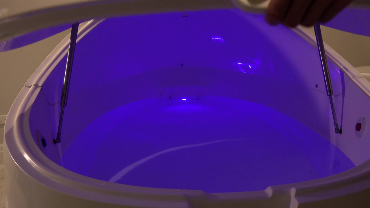 A moving wide shot of a floatation spa being opened revealing a lit pool of water inside. There is a choice of two openings with different colors inside