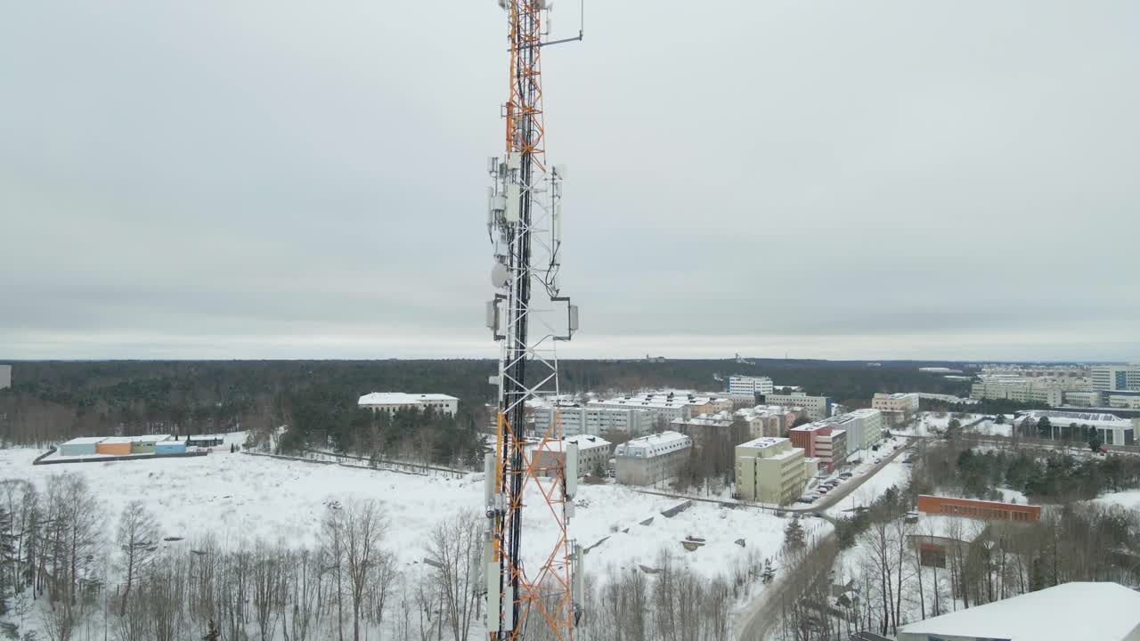 Aerial ascent in front of a frozen telecommunications tower, revealing cellular panel antennas and radio equipment coated in ice and snow above a winter city landscape