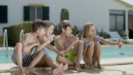 children sitting on poolside taking selfie on sunny day.