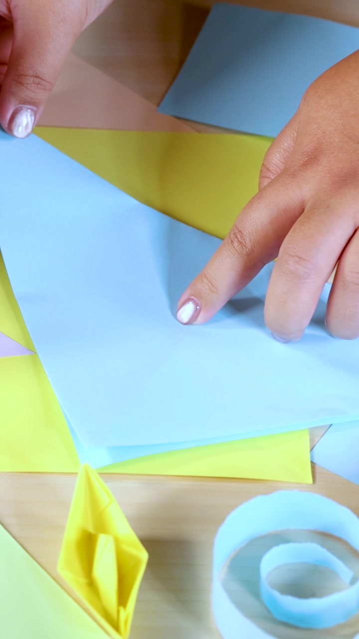 Una persona piega carta origami blu su un tavolo di legno con illuminazione brillante e materiali artigianali colorati.