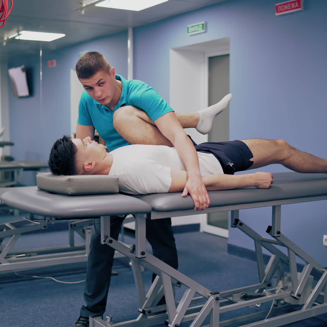Physiotherapist is leaning and stretching patient's leg a medical center. Young masseur is giving leg treatment to a client lying on a massage table. Physical therapy concept.