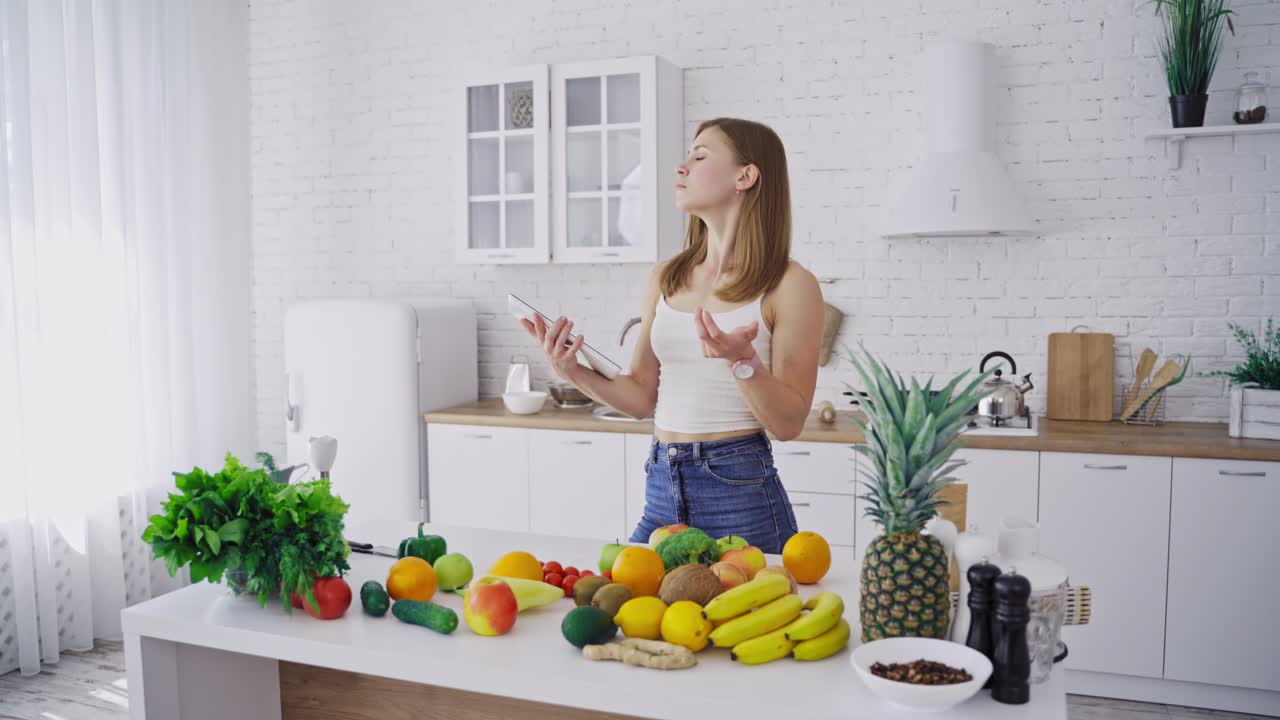Pretty woman holds tablet in the kitchen. Young female is thinking about a new recipe of organic food. Fresh fruit and vegetables on the table. Healthy eating.