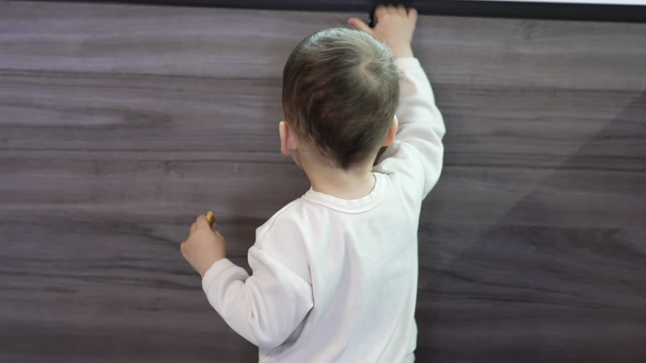 Cute toddler in white sweatshirt walks by the kitchen eating a cookie. Kid comes up to a cupboard and hold his hand on the door.