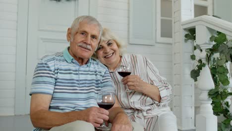 Senior-elderly-couple-drinking-wine,-embracing-in-porch-at-home-during-coronavirus-quarantine