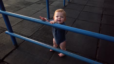 un niño pequeño feliz está solo en una alfombra de juegos de goma apoyado contra el gimnasio de la jungla azul mirando por encima de los bares