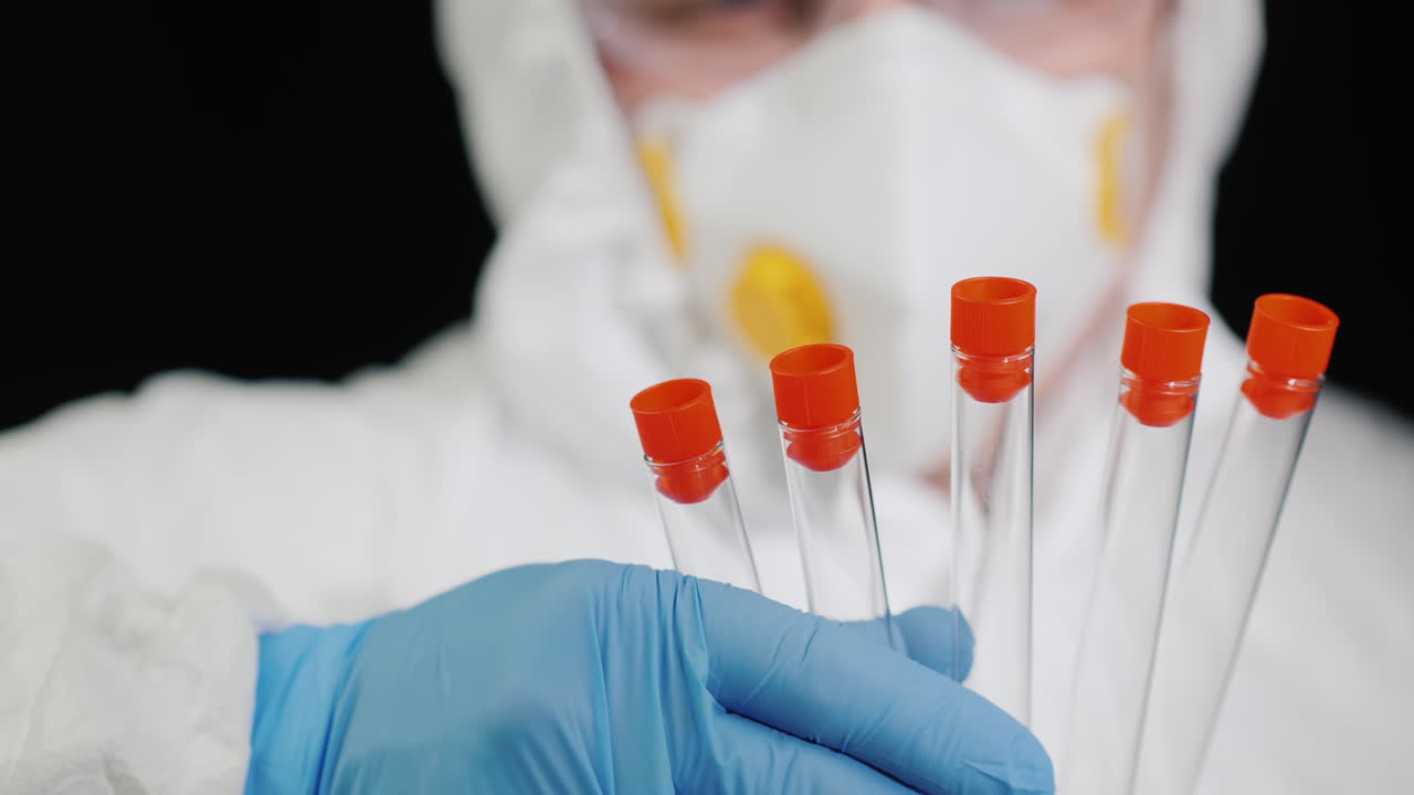 A man in protective clothing and a respirator holds several test tubes