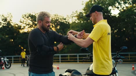 Driving-instructor-in-yellow-t-shirt-helping-a-biker-with-a-beard-to-put-on-elbow-protection.-Protective-clothing-as-an-important-part-of-a-motorcycle-trip