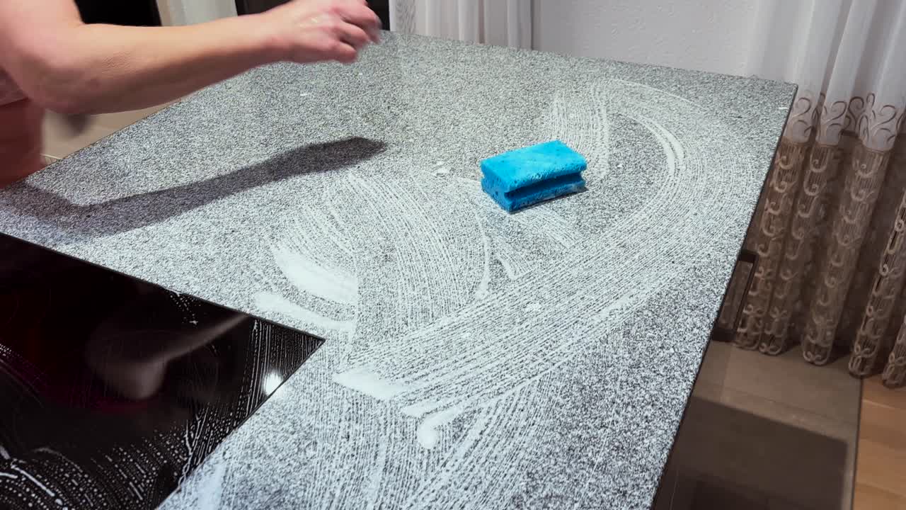 A person wipes and cleans a kitchen countertop with a sponge, focusing on hygiene, cleanliness, and daily household maintenance