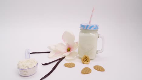 Tiro-Giratorio-Lento-De-Frijoles-De-Vainilla,-Leche-En-Una-Taza-De-Tarro-De-Albañil,-Hermosa-Flor-Blanca-Y-Sabrosas-Galletas-En-Forma-De-Hoja