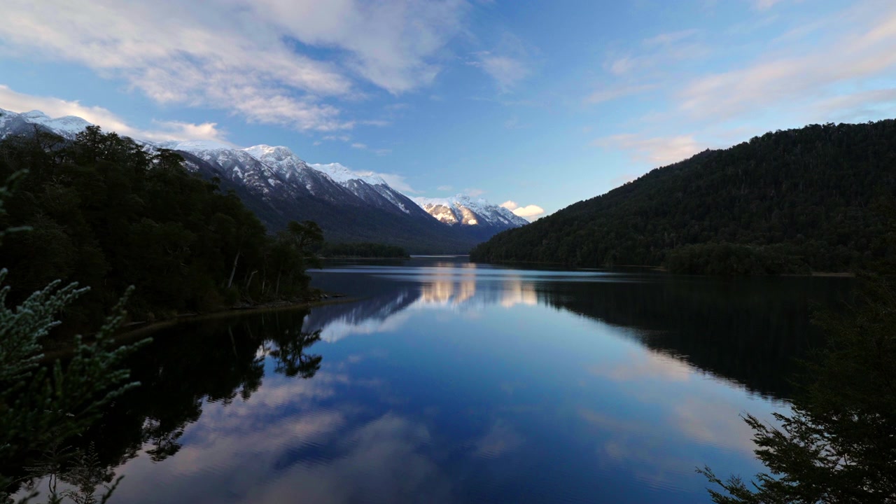 Cime innevate si specchiano nelle acque tranquille di un lago remoto della Patagonia al tramonto. Ideale per documentari naturalistici o servizi di viaggio che mostrano paesaggi sereni.