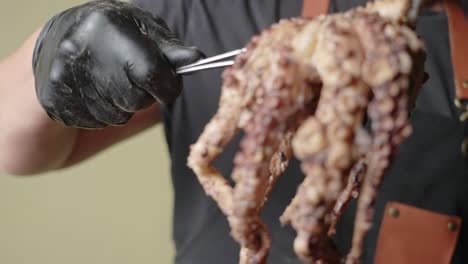un chef masculino con guantes negros, sostiene un pulpo medio cocido con una pinza de una tabla de cortar, disparo en cámara lenta
