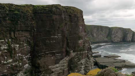 the camera slowly tilts downwards to reveal a breathtaking view of a seabird colony with dramatic sea cliffs as turquoise green waves crash against the base of cliff