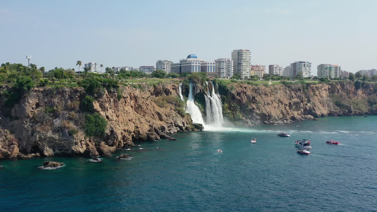 aerial drone of the waters of Lower Duden Waterfall drop off a rocky cliff directly into the Mediterranean Sea in Antalya Turkey with large buildings overhead and local tourist boats viewing below
