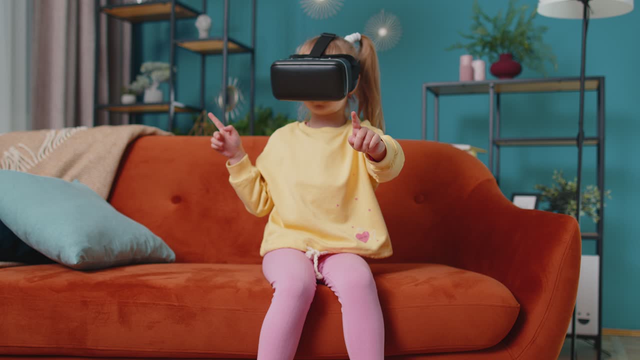 Toddler girl sitting on home sofa using virtual reality headset helmet app to play simulation game