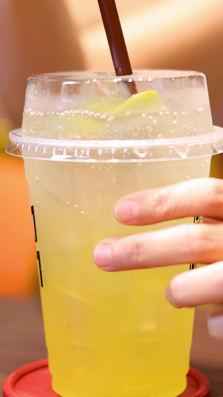 A hand inserts and stirs a straw in a plastic cup of lemon soda with ice and lime, under warm indoor lighting on a wooden table