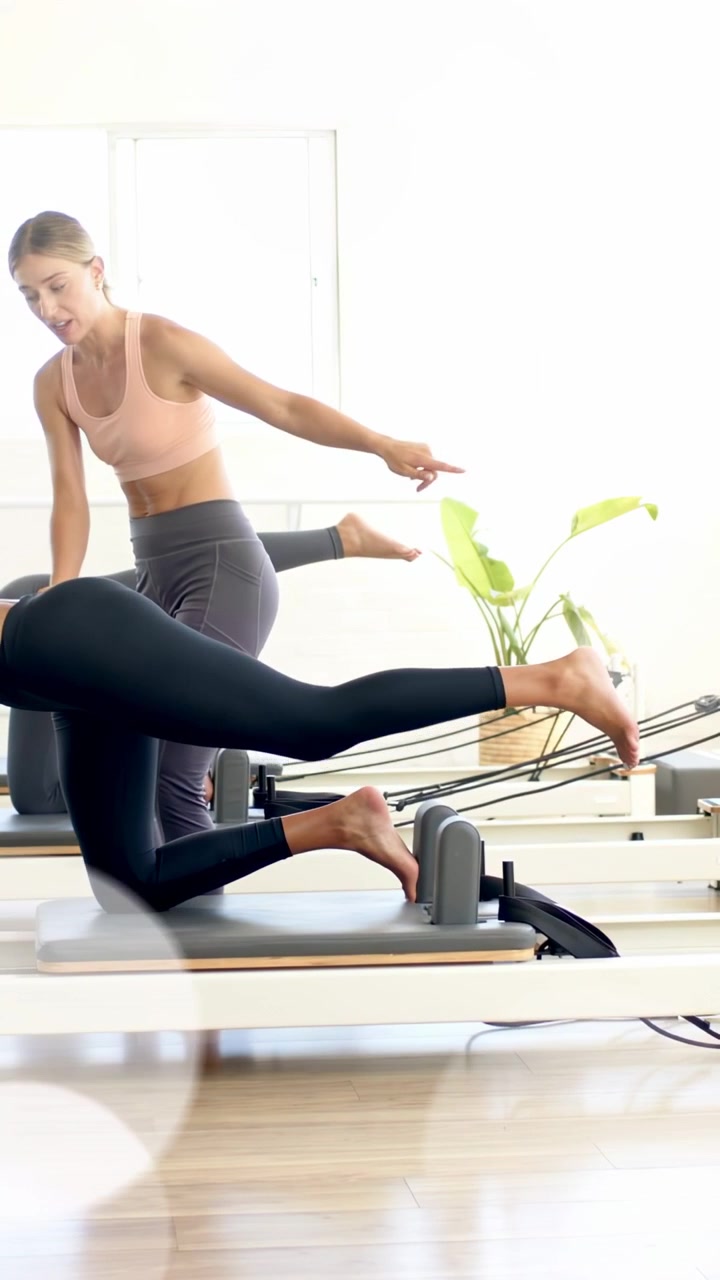 Vertical video: Coach cueing women in Pilates, extending legs for core, bokeh highlighting motion