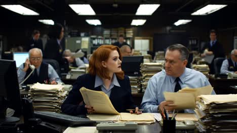 in a bustling office filled with stacks of documents, a focused woman reviews papers while a man beside her works diligently, embodying the diligence of the work environment