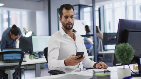 Hombre-De-Negocios-De-Raza-Mixta-Sentado-En-El-Escritorio-Usando-Computadora-Y-Teléfono-Inteligente-En-Una-Oficina-Ocupada