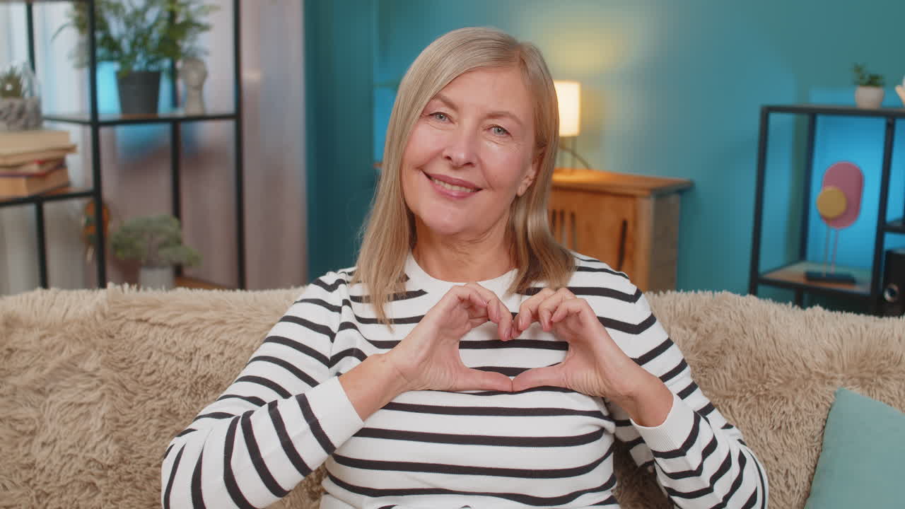 Mature woman making heart gesture on sofa expressing love and gratitude showing affection at home