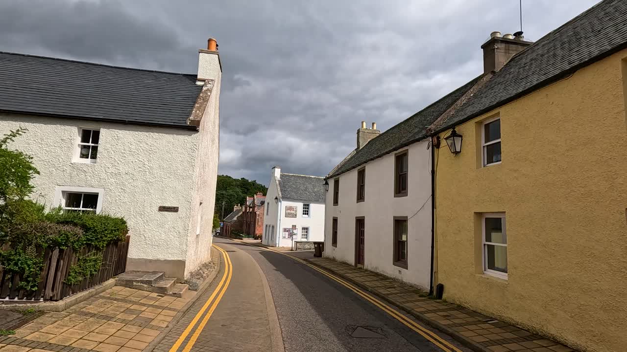 A steady daytime walk down a quiet Cromarty street, lined with pastel traditional houses under overcast skies, captured in smooth forward camera movement
