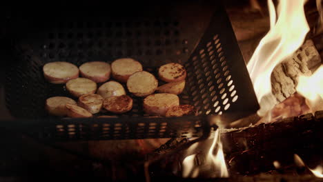 slices of potatoes are fried in a frying pan in the grill, next to the frying pan flames and hot coals