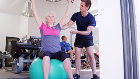 Senior-Woman-Exercising-On-Swiss-Ball-With-Weights-Being-Encouraged-By-Personal-Trainer-In-Gym