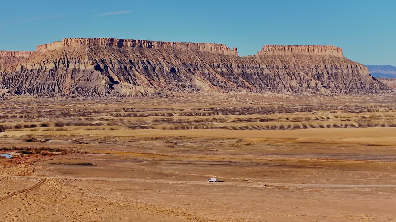 Un veicolo si dirige verso una cresta desertica e mesas che si estendono attraverso l'arido paesaggio dello Utah sotto un cielo sereno, ripresa con drone.