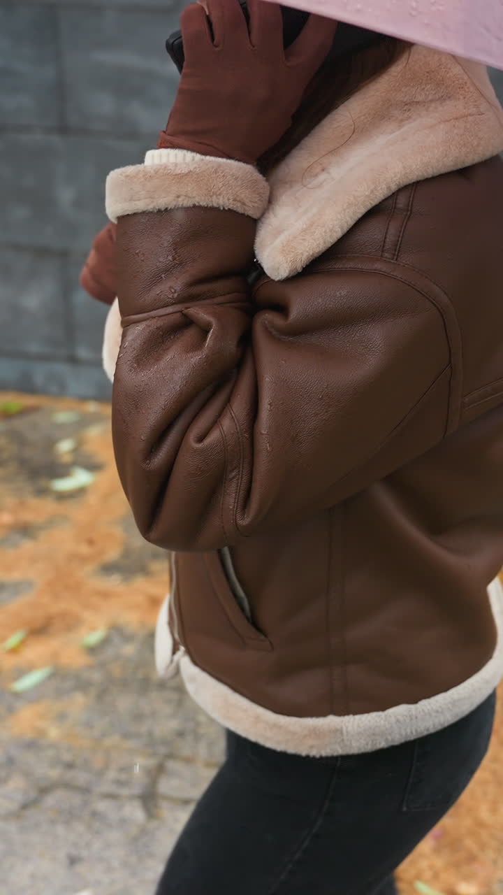 Up shot of young girl smiling on phone call holding umbrella, wearing knit cap, brown shearling jacket, black trousers, walking down stone steps during light snowfall with colorful autumn leaves