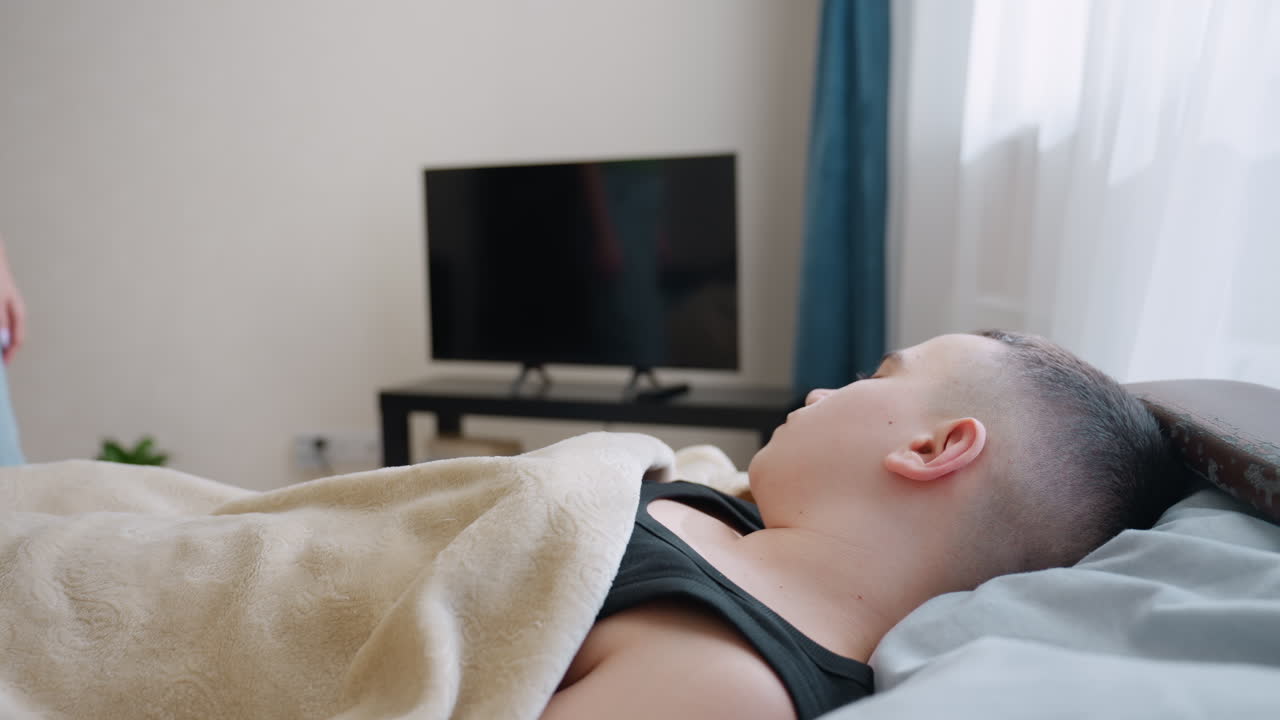 Caring mother squats beside bed gently taps son on nose to wake him up as he lies under blanket with television in background showing tender parenting moment of love