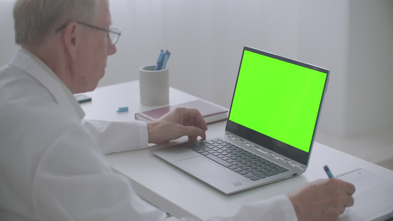 un anciano médico está leyendo un artículo en internet mirando la pantalla de la tecla de croma de la computadora portátil y escribiendo