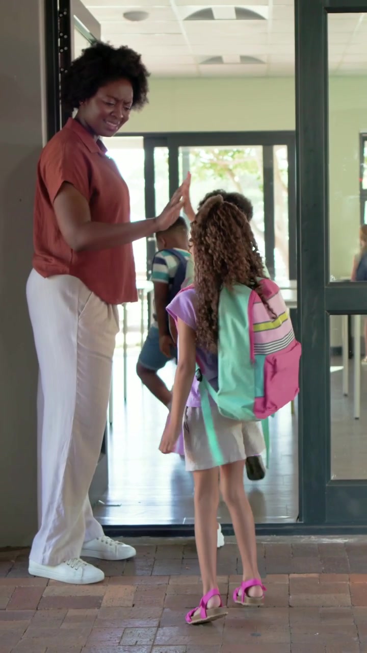 Vertical video: Greeting teacher in rust, white sneakers hi-fiving kids enter with bags, copy space