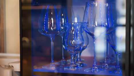 close up of elegant wine glasses arranged inside cabinet under soft blue lighting with reflections creating vibrant highlights on glass surfaces, capturing modern bar ambiance