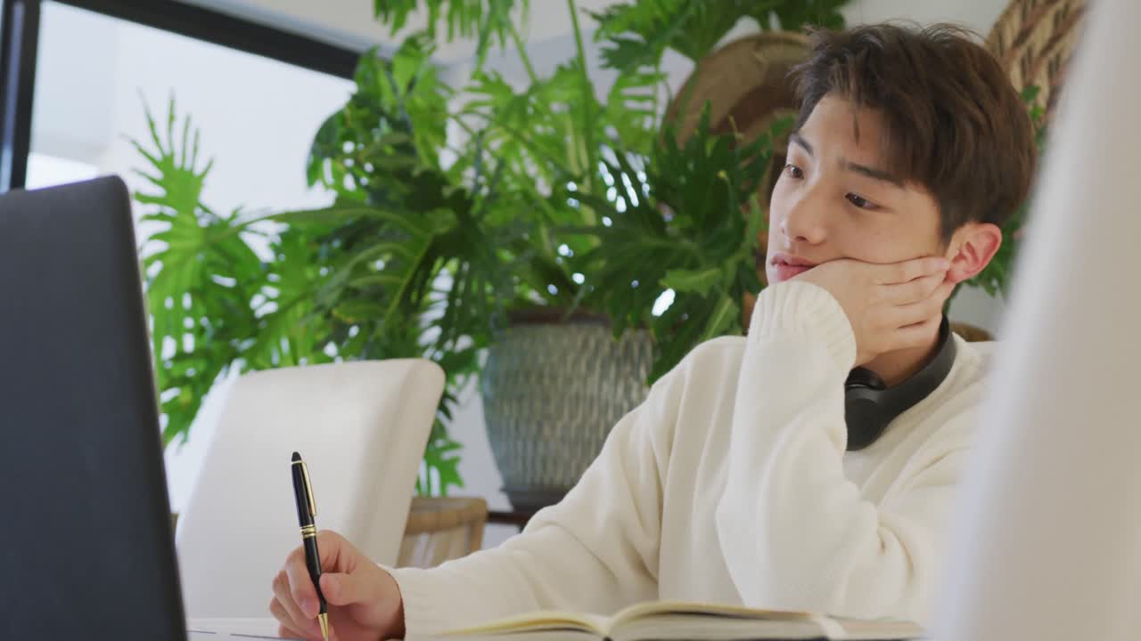 헤드폰을 착용한 아시아 남성 십대 청소년이 거실에서 노트북을 배우고 사용하고 있습니다.