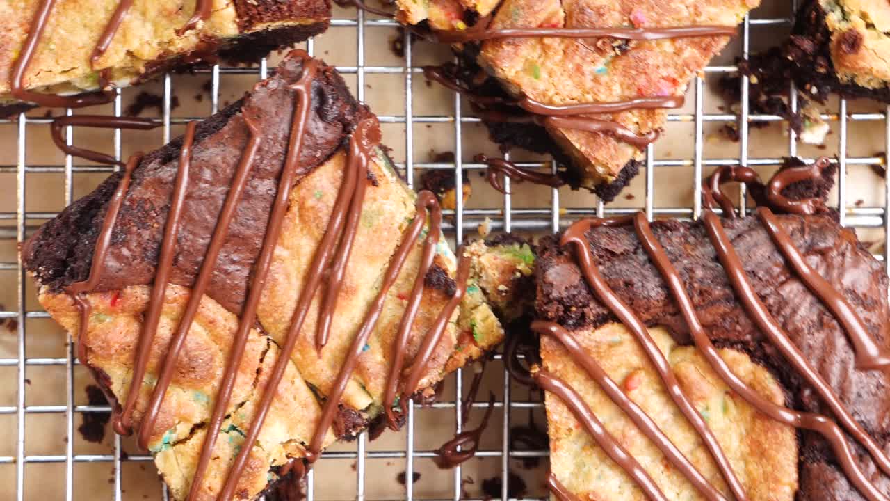 Baking chocolate brownies with added confetti sugar cookies, called a Brookie. Close up and panning across brownies drizzled with melted chocolate on a wire rack. In the kitchen making a dessert
