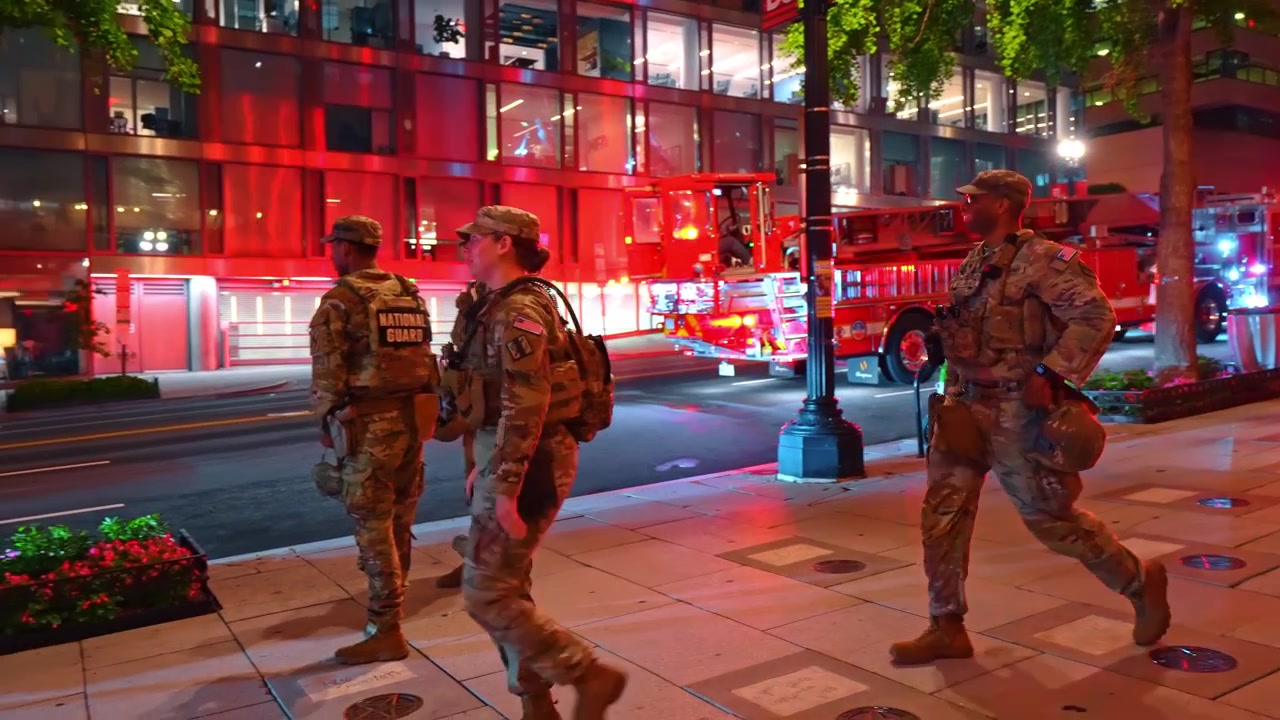 Washington, USA, 12 June 2026: National Guard troops walking along a city sidewalk at night, Washington DC. Four soldiers in camouflage pass storefronts under red ambient glow.