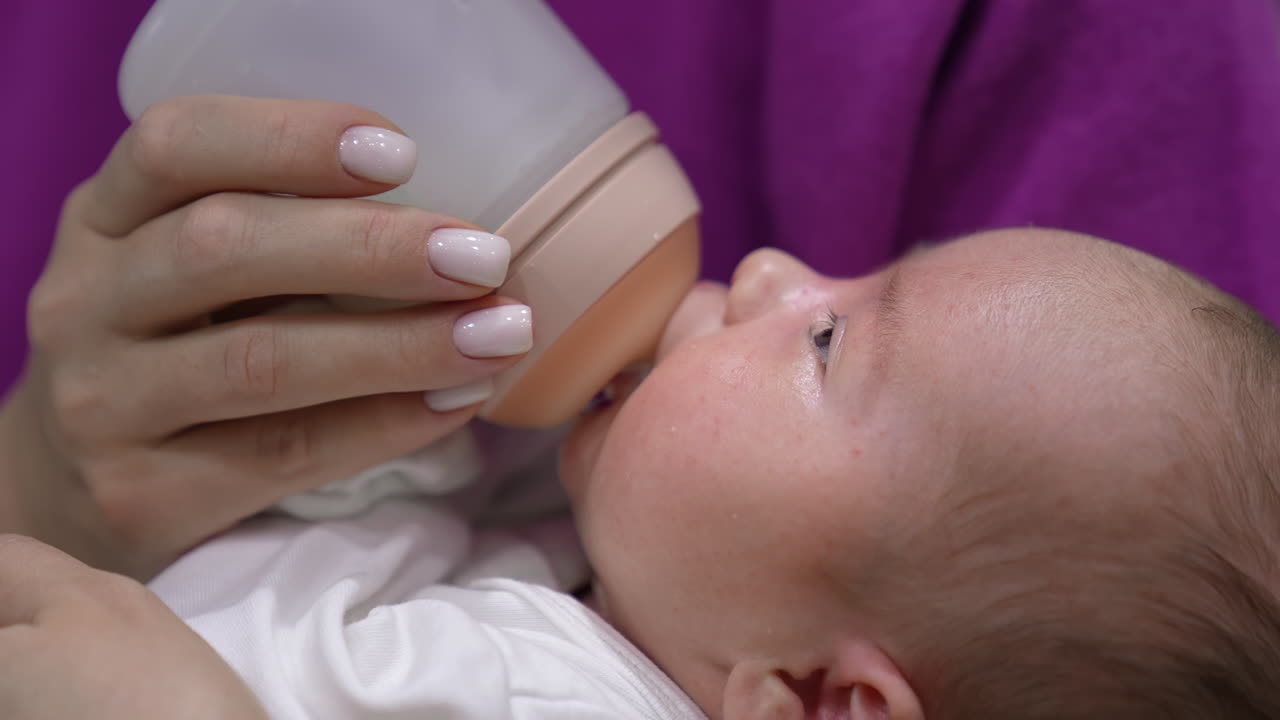 Infant babe is being fed by his mum from a bottle. Kid suckling milk from the bottle and slowly falling asleep. Close up.
