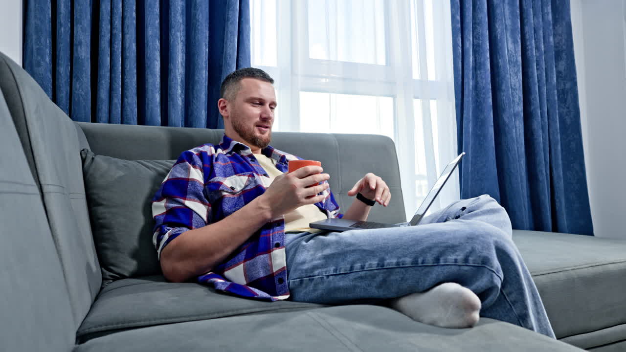 Man sits on gray couch and drinks coffee while using laptop in a bright room with blue curtains during the afternoon