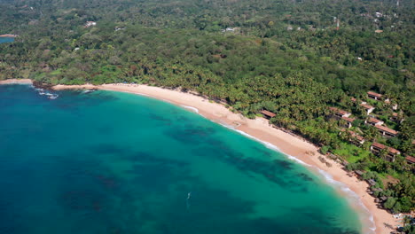 slow drone push-in with a gentle pan, starting from colorful beach umbrellas and revealing a tropical palm shoreline and turquoise sea at silent beach, sri lanka.