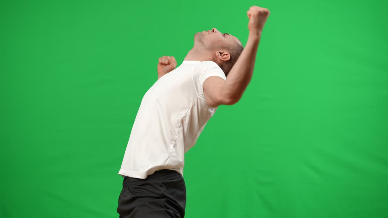 Live camera follows Caucasian football player standing up at chromakey background gesturing victory. Satisfied skilled young sportsman rejoicing goal on green screen. Achievement and win concept.