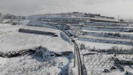 Statische-Luftaufnahme-Von-Lastwagen,-Die-Die-Straße-Im-Schnee-Eines-Terrassenförmig-Angelegten-Weinbergs-Und-Eines-Dorfes-In-Israel-Hinauffahren