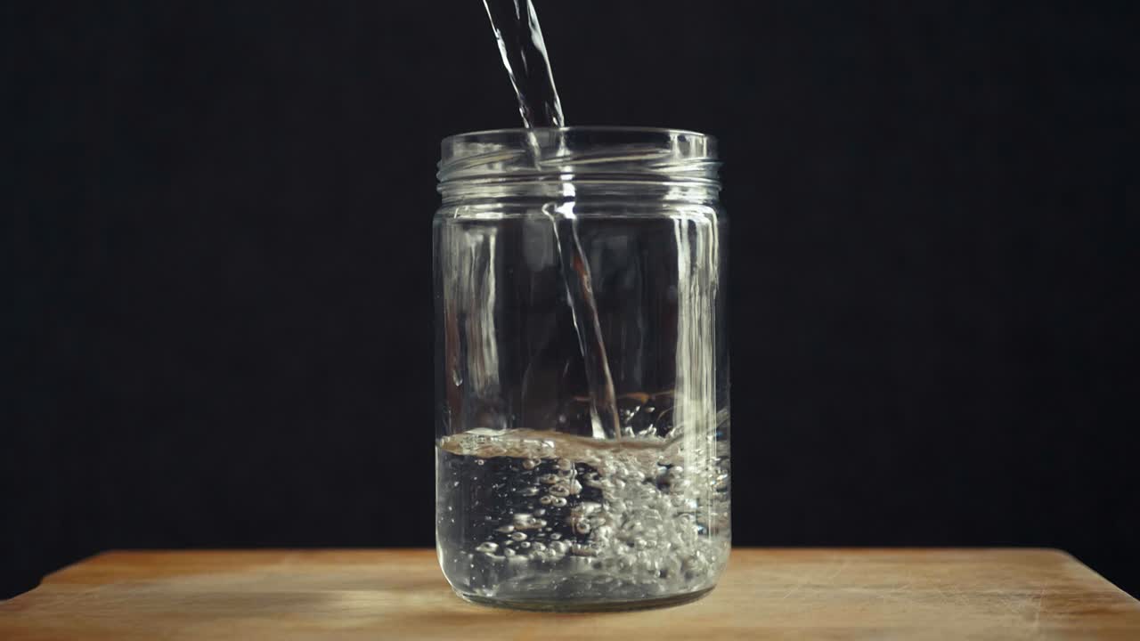 Water being poured into a clear glass jar on a wooden surface against a dark background