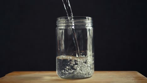 water being poured into a clear glass jar on a wooden surface against a dark background