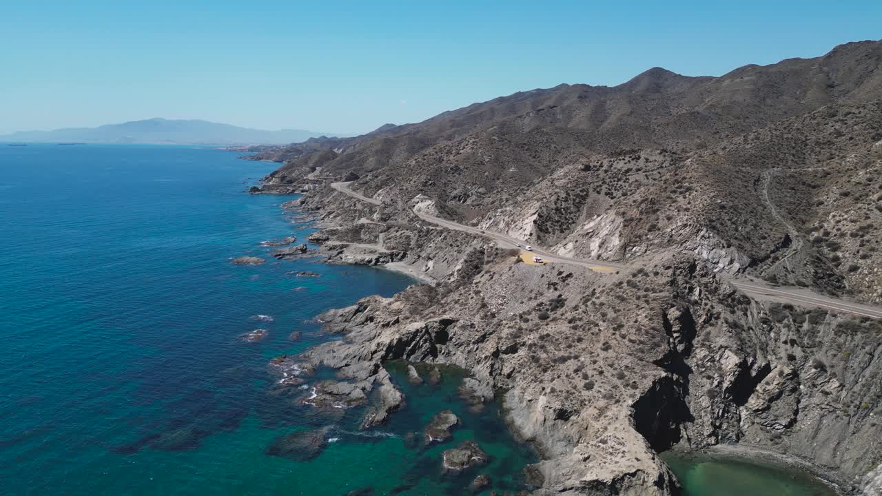 Slow aerial pullback showing a campervan parked along the rugged seaside cliffs of the Almería coast in southern Spain, revealing more of the winding coastal road and rocky Mediterranean landscape
