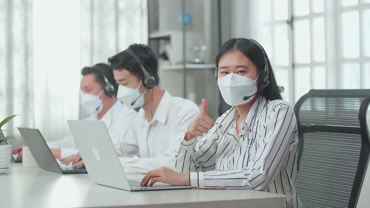 A Woman Of Three Asian Call Center Agents Wearing Headset And Mask Looking At Camera And Thumbs Up While Two Of Her Colleagues Are Speaking With Customer At The Office