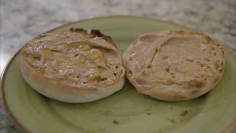 Einen-Gerösteten-Englischen-Muffin-Mit-Erdnussbutter-Auf-Einem-Grünen-Teller-Auf-Einer-Marmorarbeitsplatte-Mit-Honig-Beträufeln-Und-Verteilen