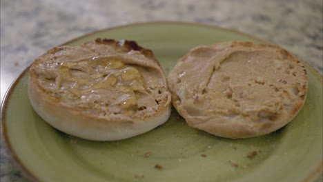 llovizna y esparce miel en un muffin inglés tostado con mantequilla de maní en un plato verde en una encimera de mármol